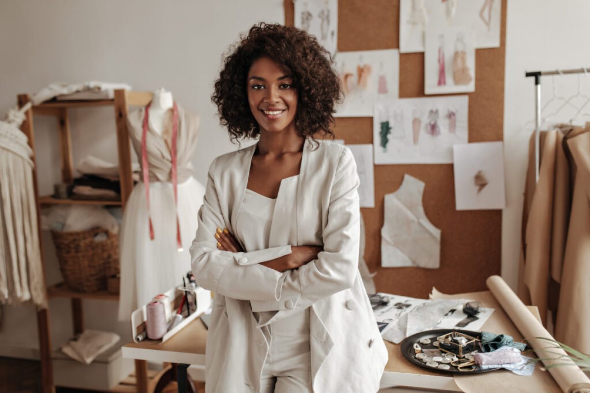 Beautiful curly brunette dark-skinned fashion designer poses in office, leans on table. Young lady